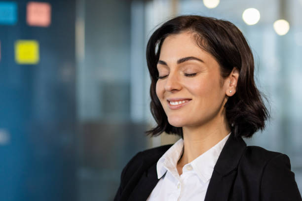 Professional woman meditating and finding peace in an office environment with a serene expression and eyes closed. Embracing mindfulness during a busy workday for mental wellness.