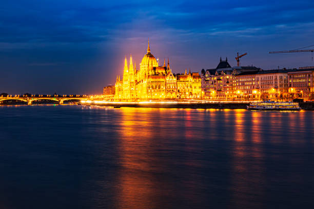 Budapest, Hungary, April 2025The Hungarian Parliament Building stands prominently along the Danube River in Budapest, Hungary, captured at night with warm golden illumination accentuating its Gothic Revival architecture, including tall spires, intricate facades, and a central dome. The structure's lights reflect softly on the calm river water, creating a symmetrical glow against the dark night sky. No people are visible in this wide-angle view of the historic landmark. The image evokes a sense of grandeur and timeless elegance, suitable for commercial uses such as travel articles on European capitals, tourism campaigns promoting Budapest, editorial pieces on Hungarian history and architecture, book covers for travel guides, or media content illustrating nighttime cityscapes and government buildings.The Hungarian Parliament Building stands prominently along the Danube River in Budapest, Hungary, captured at night with warm golden illumination accentuating its Gothic Revival architecture, including tall spires, intricate facades, and a central dome. The structure's lights reflect softly on the calm river water, creating a symmetrical glow against the dark night sky. No people are visible in this wide-angle view of the historic landmark. The image evokes a sense of grandeur and timeless elegance, suitable for commercial uses such as travel articles on European capitals, tourism campaigns promoting Budapest, editorial pieces on Hungarian history and architecture, book covers for travel guides, or media content illustrating nighttime cityscapes and government buildings.