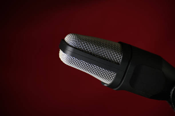Close-up of a studio microphone against a dark red background, featuring a metal grille and black body, perfect for podcasts or music recording.
