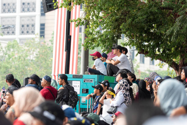 Putrajaya, Malaysia - August 31, 2024: Spectators watch attentively from under trees, some perched on elevated platforms, at a large public gathering, with a backdrop of buildings and greenery