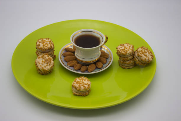 Elegant coffee setup with a porcelain cup, whole almonds, and stacks of cream-filled cookies on a vibrant green plate. A charming display of treats for a cozy moment.
