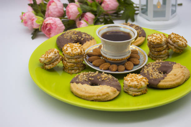 Porcelain cup of coffee surrounded by almonds and layered cookies on a vibrant green plate. Romantic backdrop with pink roses and a white candle lantern.