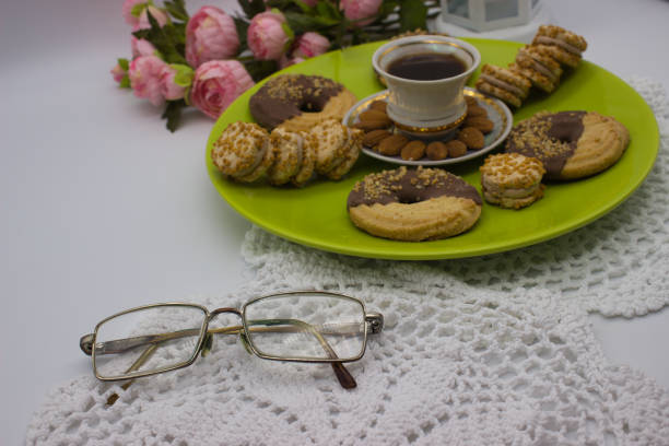 A warm coffee with almond garnish, assorted cookies on a green plate, and vintage glasses resting on a delicate crochet cloth. Pink roses add a soft touch.