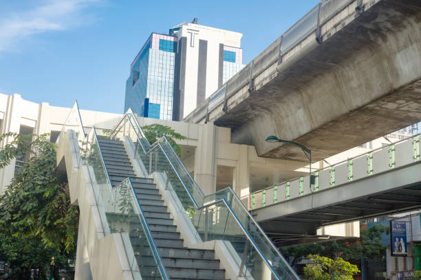 Steps to BTS skytrain and footbridge at Sukhumvit Road in Bangkok downtown. At building tower in background is a huge T. At right side are stores and banners at opposite side of street