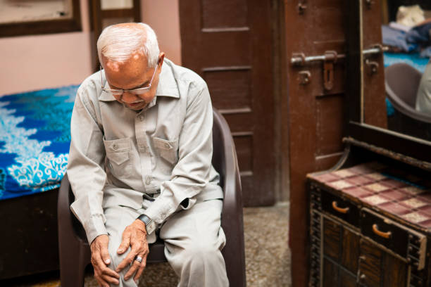 Senior Indian man sitting on the chair and suffering from knee joint pain at home.