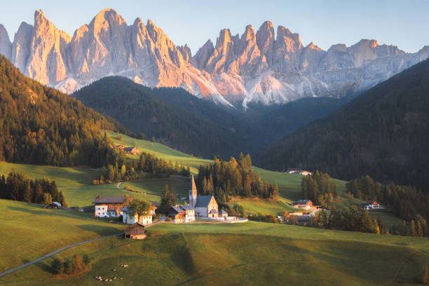 View of charming village Villnöss and Church of St. Magdalena in Val di Funes, surrounded by lush meadows and set against the majestic peaks of the Dolomites, bathed in warm sunset light, South Tyrol, Italy.