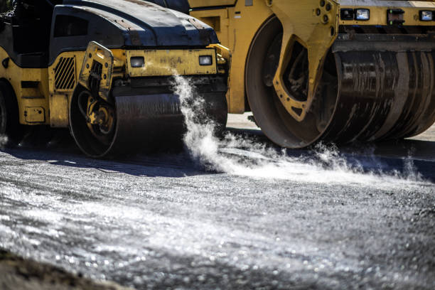 Steam rises from asphalt as heavy rollers compact a newly laid road in a construction zone during the day Heavy machinery works diligently to compact asphalt in a construction area, creating steam that rises from the hot surface under bright sunlight. hot mix asphalt stock pictures, royalty-free photos & images