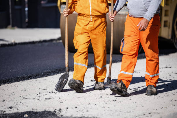 Two construction workers in orange overalls using tools to prepare a road surface for asphalt paving during daylight
