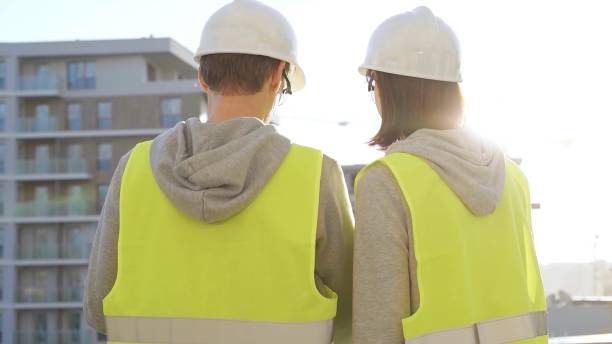 istock Two architects wearing safety hard hats and vests holding blueprint and discussing construction site project at sunrise, back view. Architecture, engineering and teamwork concept 2181236442