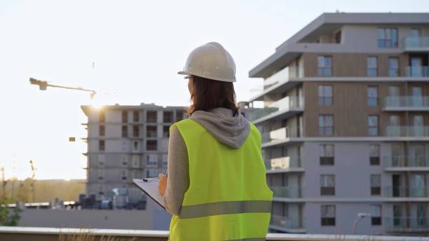 istock Female construction engineer with a hard hat and safety vest is writing on a clipboard while inspecting a construction site in the evening at sunset 2181237243