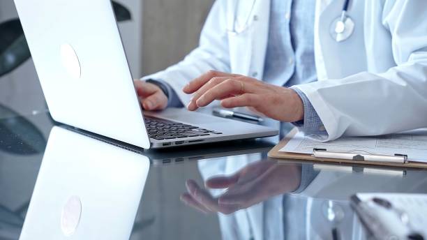 istock Doctor man with blue shirt and medical coat is using laptop computer in clinic office while sitting at the glass desk. Medicine concept 2181240227