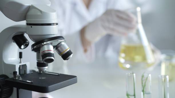 istock Microscope is near a scientist, wearing a lab coat and white protective gloves, and holding beaker with a yellow oily liquid in laboratory setting, close up. Medicine and science concept 2181254367