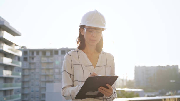 istock Woman construction engineer wearing white checked blouse and hard hat is making notes on a clipboard while inspecting a building site at sunset, front view. Architecture and engineering concepts 2181257242