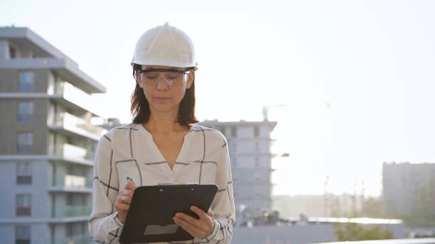 istock Woman construction engineer wearing white checked blouse and hard hat is making notes on a clipboard while inspecting a building site at sunset, front view. Architecture and engineering concepts 2181257749