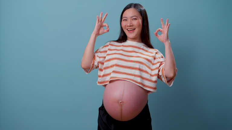 Asian pregnant woman happy confident showing OK sign by hand standing isolated over blue background.