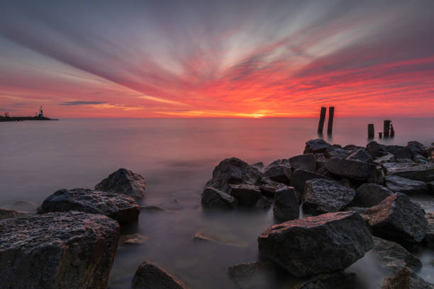Quietly enjoying the serene landscape by the IJsselmeer, where basalt blocks, wooden poles, and a green lighthouse are surrounded by a spectacular red sky. The stones and poles in the foreground, along with the lighthouse on the horizon marking the entrance to the harbor, create a beautiful sense of depth. The long exposure adds tranquility to the photo. A grand way to enjoy the Netherlands as a land of water.The IJsselmeer is part of the IJsselmeer region and was created by closing off part of the Zuiderzee. The Afsluitdijk was completed on May 28, 1932, and the official name change of the enclosed part of the Zuiderzee took place on September 20, 1932. The lake was named after the river IJssel, which flows into it.