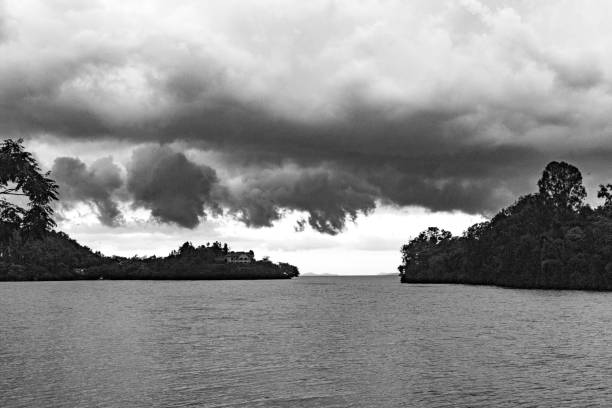 A dramatic black-and-white photograph capturing the intensity of nature as storm clouds gather over a serene lake. The dark, textured clouds create a powerful contrast with the calm water below, framed by lush forested hills on both sides. This image evokes a sense of tranquility and anticipation, highlighting the beauty and unpredictability of natural landscapes.
