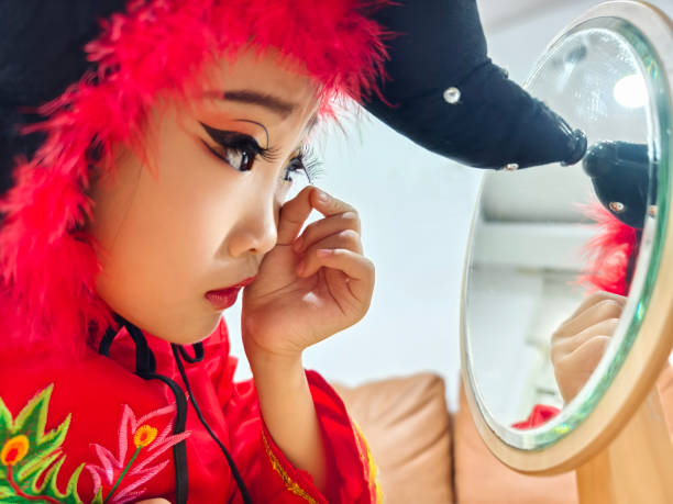 An Asian girl puts on heavy makeup (stage makeup) before performing on stage. She checks whether her makeup is complete in front of the mirror, and adds lipstick, eyebrow line, and powder herself. She was wearing the outfit she was going to go on stage, and her whole body was red. , looks cheerful and has the atmosphere of the Lunar New Year