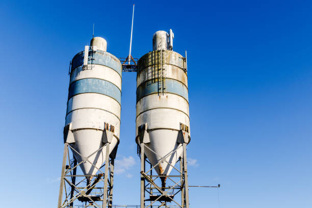 Two large industrial silos against a clear blue sky. The silos are cylindrical with a conical bottom and a metal frame. They are painted white with blue horizontal stripes and show signs of wear and tear. The silos are connected by a metal walkway