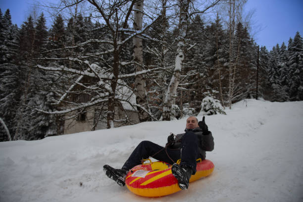 istock A man with a smile on his face and a raised hand enjoys a winter day while going down a snowy slope with a rubber sled 2181371201