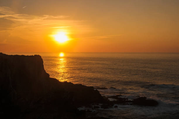 Sunset in Portugal, with an orange sun hiding on the horizon. Backlighting a rock on the cliff. The waters of the sea are calm. Copy Space