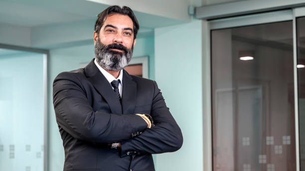 istock A middle-aged, dark-haired, bearded businessman stands in an empty office environment with a blurred background, arms crossed, smiling as he looks at the camera 2181390263