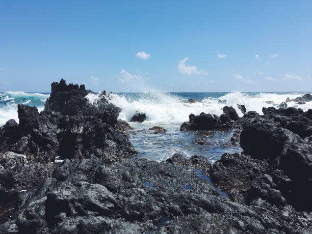 A striking view of ocean waves crashing against rugged black volcanic rocks under a clear blue Hawaiian sky. This image captures the dynamic energy of Hawaii's coastline, with powerful waves meeting the rocky shore, surrounded by the serene beauty of tropical skies and the endless Pacific Ocean. Ideal for showcasing the raw and natural beauty of Hawaiian seascapes.