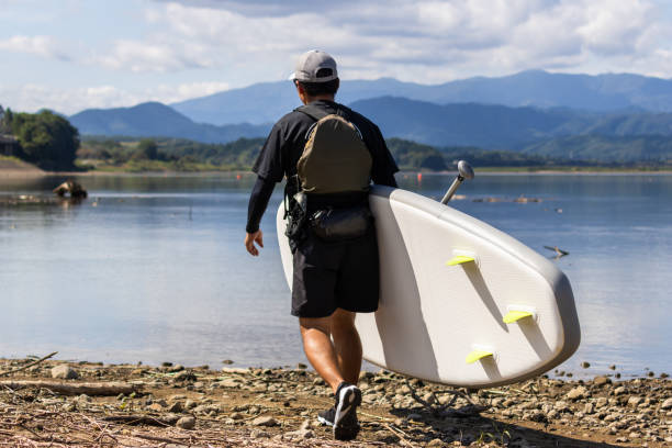 Rear view of a man carrying his Stand up paddle board to a tranquil lake.