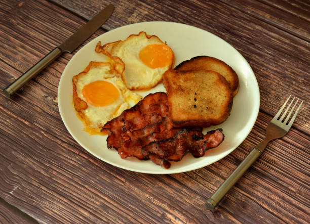 A close-up shot of crispy bacon sizzling over a cooking pan on a portable mini stove at a campsite. The smoky aroma fills the air as the bacon reaches golden perfection, with the rustic charm of the outdoors in the background.