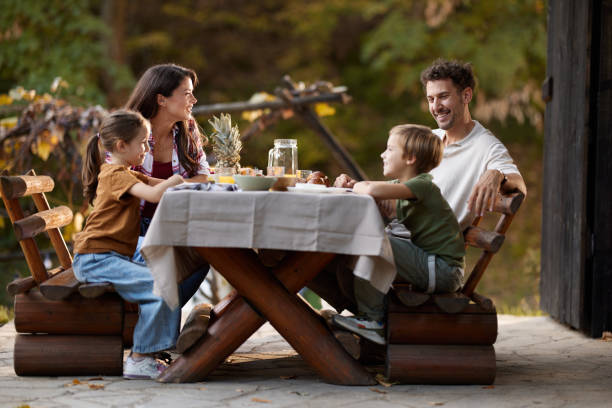 Happy family talking during a meal at the table on a patio. Happy family enjoying in conversation while having a meal at picnic table on a porch. Focus is on parents. fall picnic table stock pictures, royalty-free photos & images