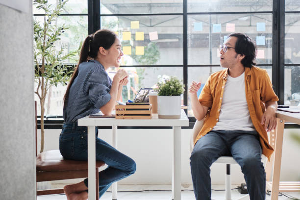 istock Two Asian workers at working desks near windows talk together. 2181455496