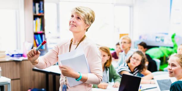 istock Woman teacher in a classroom holding a marker teaching with attentive students. 2181456412
