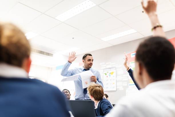 istock Man teacher engages students in a classroom, holding papers and raising a hand. 2181457542