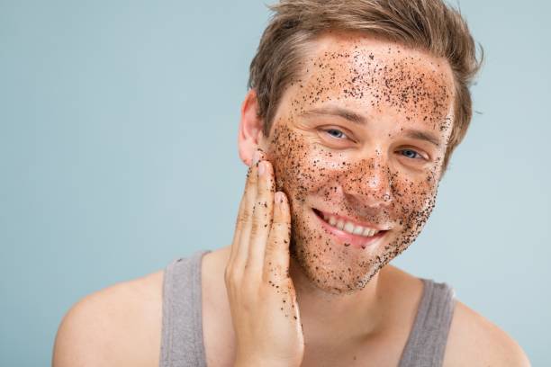 A person applying a facial scrub, smiling, with a light blue background.