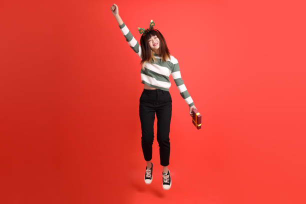 Asian woman wearing Christmas or winter clothes with Santa cap, holding a pile of presents in her hands with gesture of  Jumping isolated on red background. Xmas, Santa Claus.