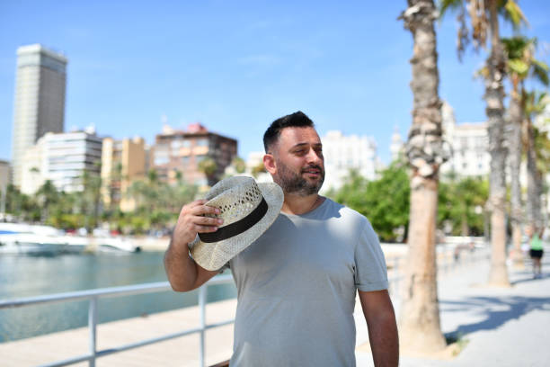 Male Adult Cooling Off With Straw Hat While Walking and Exploring Harbor With Palm Trees in Background