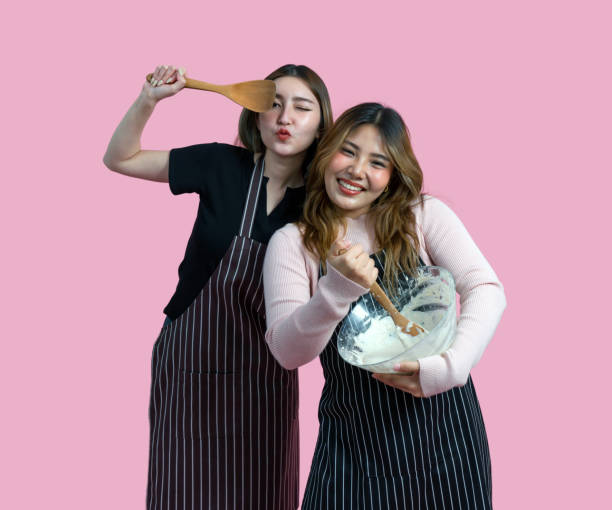 istock Two women are having fun baking together. They are wearing apron and smiling while mixing ingredients with a wooden spoon. Portrait on pink background with studio light. Isolated 2181502841