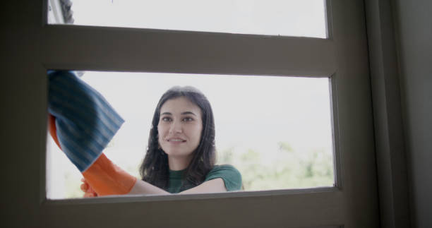 A charming woman is cleaning her home, using cloths and spraying disinfectant while wiping the glass on the window during the day.