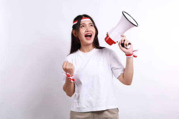 istock Excited Asian woman wearing red white flag ribbon, shouting using a megaphone while clenching hand, isolated on a white background, celebrate Indonesia independence day 2181515001