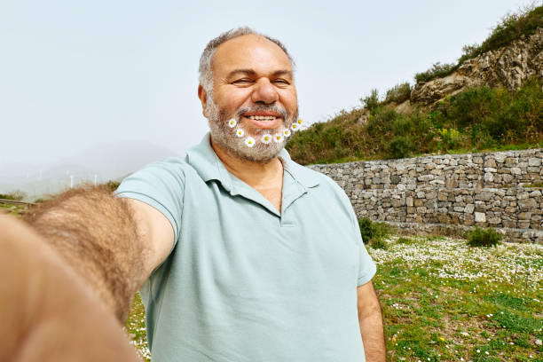 Laughing mature man with with daisies in his beard making selfie.