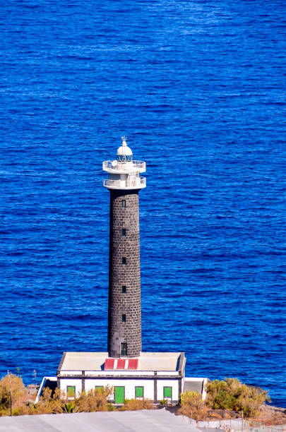 A lighthouse is on a rocky shoreline next to the ocean. The lighthouse is tall and has a red roof. The ocean is calm and blue