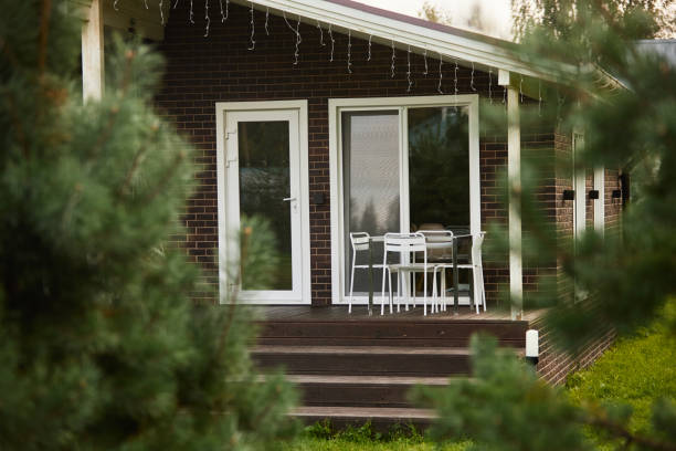 This photo is of a the front pouch and front door and front patio of a home. and there is an american flag waving in the wind. there is also the front yard with flowers and green grass. the lighting in natural outdoor sunlight in the evening. The house is a modern house with a glass front door and windows. and there is a brick house in the background. 