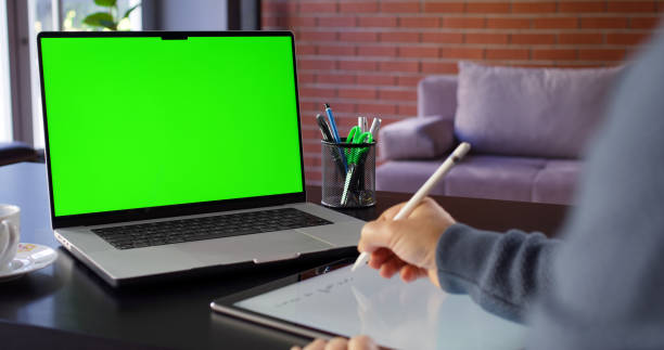 istock Man Using Laptop Computer With A Chroma Key Green Screen In The Office And Taking Notes On Digital Tablet Using Digitized Pen 2181573047