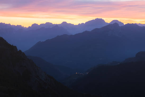 Twilight view of the Italian Dolomites from Giau Pass, with a colorful sunset sky illuminating the rugged mountains and a distant village Selva di Cadore in the valley.
