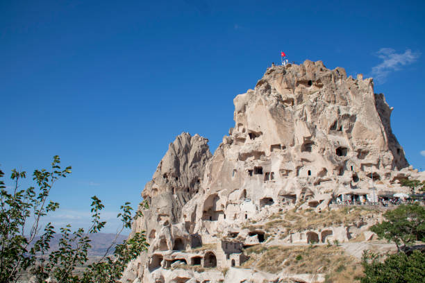 A significant and impressive rock formation stands tall, featuring a flag proudly waving at the top of it, symbolizing a journey or adventure. A rock castle with cave passages inside, located in the city of Uchisar in the Cappadocia region