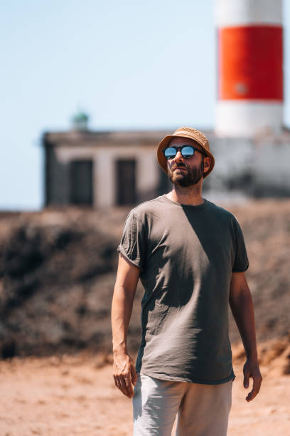 On a sunny day, a man enjoys the beach while dressed in casual clothes. The lighthouse looms in the background, its vibrant colors contrasting with the sandy landscape.