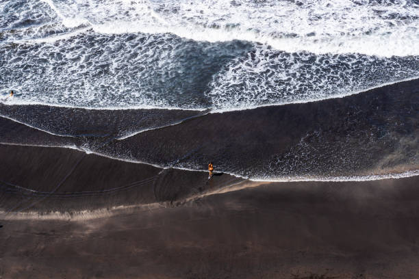 istock A solitary figure walks along a black sand beach at high tide, waves crashing 2181599755