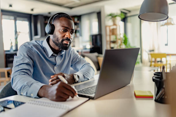 istock African-American man wearing headphones, taking notes while working on a laptop from home, focused and engaged in a modern workspace. 2181600834