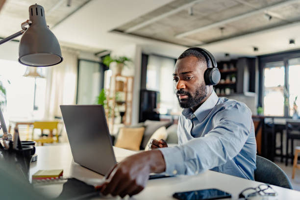 istock African-American man wearing headphones, taking notes while working on a laptop from home, focused and engaged in a modern workspace. 2181600980