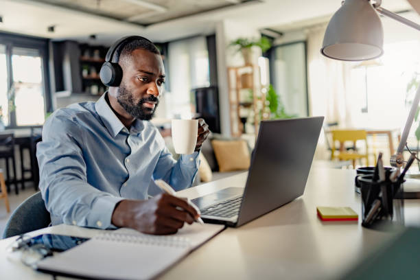 istock African-American man wearing headphones, taking notes while working on a laptop from home, focused and engaged in a modern workspace. 2181601060